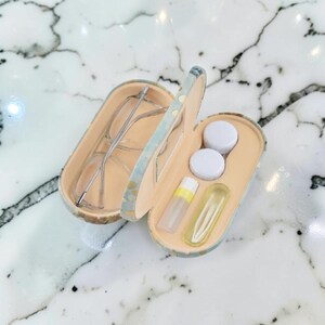 Könnte beinhalten: Geöffnete Brillenetui mit einer Brille mit silbernem Rahmen. Das Etui ist beige mit blau-grünen Musterrändern. Die andere Seite des Etuis enthält zwei Behälter mit weißem Deckel, eine kleine Flasche und einen gelb getönten Behälter.