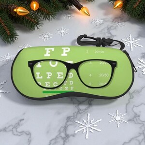 Op de afbeelding: Een limoengroene brillenkoker met een zwart brillenmontuurontwerp over een klassieke oogtestkaart. De koker heeft een zwarte rits en een zwarte clip. De achtergrond bevat kerstversieringen.