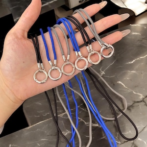 以下が含まれることがあります： 黒、青、グレーのストラップのコレクション。各ストラップは、銀色の金属リングとそれに合わせたコードが特徴です。コードは織り込まれた素材で作られています。ストラップは大理石のような表面に表示されています。