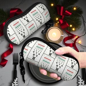 Op de afbeelding: Twee witte brillenkokers met een zwarte rits en een patroon van oogkaarten. De kokers hebben een rood en groen lijnenontwerp. Eén koker wordt door een persoon vastgehouden. Op een tafel met een kaars en rood lint.