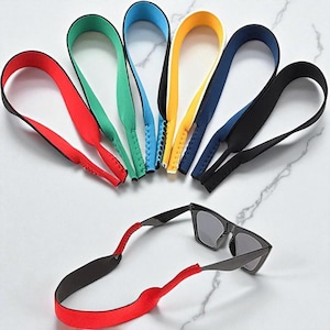 Puede incluir: Una colección de correas de gafas de sol coloridas en rojo, verde, azul, amarillo y negro. Un par de gafas de sol está sujeto a una correa roja. Las correas están hechas de un material suave y flexible.