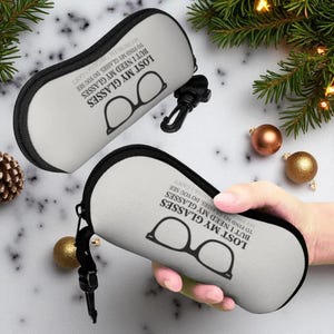 Op de afbeelding: Twee ovale brillenkokers met zwarte rand en ritsen. De kokers zijn lichtgrijs met de tekst "LOST MY GLASSES BUT I NEED MY GLASSES TO FIND MY GLASSES, DO YOU SEE MY PROBLEM BECAUSE I CAN'T" en een bril-afbeelding.