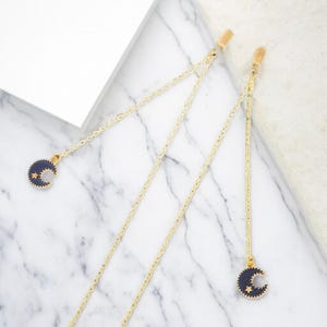 Könnte beinhalten: Zwei goldfarbene Haarspangen mit zarten Ketten. Jede Kette endet mit einem dunkelblauen Halbmond-Anhänger mit goldenen Sternen. Die Spangen sind vor einem weißen und grauen Marmorhintergrund platziert.