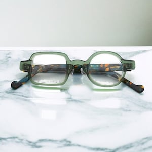 Puede incluir: Gafas únicas con montura verde. Una lente es cuadrada, la otra redonda. Las patillas y el interior de las monturas tienen un estampado carey. Las gafas están sobre una superficie de mármol blanco y gris.
