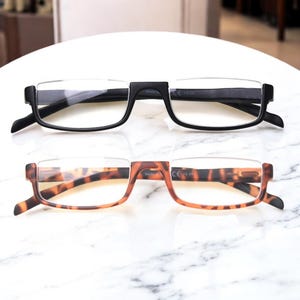 Puede incluir: Dos pares de gafas de lectura. Un par tiene montura negra y el otro un patrón carey marrón. Ambos tienen un diseño de media montura con lentes transparentes. Las gafas están sobre una superficie de mármol blanco.
