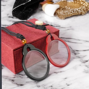 Op de afbeelding: Twee vergrootglas hangers, één zwart en één rood, met zwarte koorden en decoratieve kralen. De hangers zijn ovaal van vorm met heldere lenzen. Ze rusten op een rode doos tegen een marmeren achtergrond. Andere accessoires zijn zichtbaar.