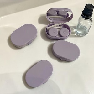May include: A selection of contact lens accessories in a pale purple colour. The set includes three closed cases, one open case with lenses and applicators, and a small glass bottle with a black cap. The open case has a mirror inside.