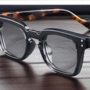 Puede incluir: Gafas de sol con montura negra y lentes grises. Las patillas presentan un diseño de carey. Las gafas tienen forma cuadrada y están sobre una superficie de madera. Diseño moderno y elegante.