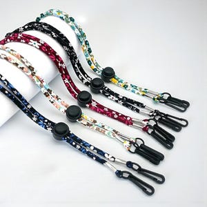 May include: Six face mask lanyards with floral patterns in various colours, including blue, white, red, and black. Each lanyard has a black adjustable toggle and black clips for attaching to a mask. The lanyards are made of fabric.