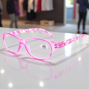 Puede incluir: Gafas de lectura rosas con un estampado floral en las varillas. Las gafas tienen una graduación de +3,00. Las monturas son de color rosa brillante y las lentes son claras.