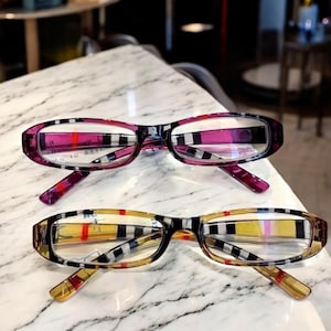 Puede incluir: Dos pares de gafas con montura rectangular. Un par tiene un patrón de cuadros rosa y negro, el otro un patrón de cuadros amarillo y negro. Ambos tienen lentes transparentes y se exhiben sobre una superficie de mármol.