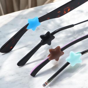 May include: Close-up of four pairs of spectacles with star-shaped silicone grips in blue, black, brown, and teal. The spectacle arms are black and tortoiseshell.