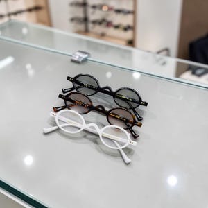Puede incluir: Tres pares de gafas de forma ovalada, en negro, marrón y blanco. Las gafas tienen lentes oscuras y se exhiben sobre una superficie reflectante. Las monturas tienen un diseño retro, de forma pequeña y redondeada.
