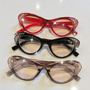 May include: Three pairs of cat-eye style eyeglasses in red, black, and taupe. Each pair features a rhinestone embellishment along the top outer edges of the frames. The lenses are clear, and the frames are made of a glossy material.