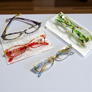 May include: Four pairs of spectacles with vibrant floral designs. The frames feature colours such as red, green, yellow, and purple. Each pair is placed on a small, textured white surface.