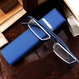 Puede incluir: Gafas de lectura azules con un estuche azul a juego. Las gafas tienen una montura rectangular con detalles plateados y el estuche tiene una banda plateada. Las gafas están sobre una superficie marrón oscuro.