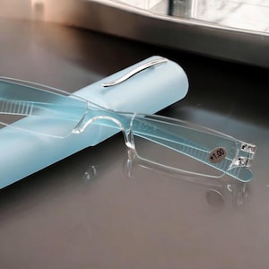 Può includere: Occhiali da lettura trasparenti con etichetta di gradazione +1,00, appoggiati su un astuccio azzurro. Gli occhiali hanno un design minimalista con montatura e aste trasparenti. L'astuccio ha una clip color argento.