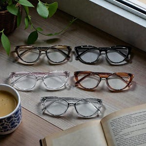 May include: An assortment of eyeglasses in various colors and styles, including clear, brown, and black frames. The glasses are arranged on a wooden surface with a cup of coffee, a plant, and an open book.