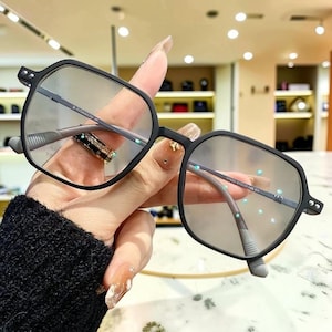Puede incluir: Gafas negras de forma geométrica con lentes transparentes. Las monturas son de color negro mate y las patillas grises claras. Las gafas están sujetas por una mano. El texto "B-TITANIUM IP" es visible en la montura.