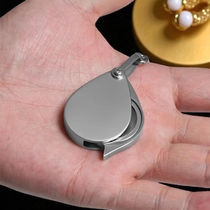 May include: A silver-colored, teardrop-shaped magnifying glass with a hinged handle. The lens is partially visible, and the design is sleek and modern. The magnifying glass is held in an open palm.