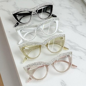 Puede incluir: Cuatro pares de gafas de ojo de gato con adornos de pedrería. Las monturas son negras, blancas, amarillas y rosas. Las gafas están dispuestas en fila, mostrando sus diferentes colores y diseños.