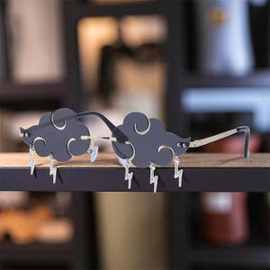 May include: Cloud-shaped sunglasses with a dark gray frame and silver lightning bolt charms. The lenses are dark, and the frame has a gold-colored bridge and black temples. The sunglasses are displayed on a wooden surface.