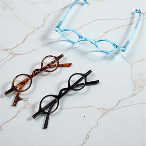 May include: Three pairs of spectacles in different colours. One pair is light blue, another is tortoiseshell, and the third is black. The frames have a unique wavy design. The lenses are round and appear to be reading glasses.