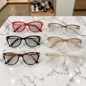 Puede incluir: Seis pares de gafas con varios colores y estilos de montura se muestran sobre una superficie de mármol blanco. Las monturas incluyen opciones rojas, negras, marrones y transparentes, con detalles dorados. Las lentes son transparentes.