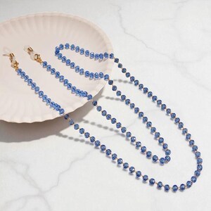 May include: A blue beaded glasses chain with gold-coloured metal connectors. The chain is made of small, faceted blue beads. The chain is resting on a white, scalloped plate.