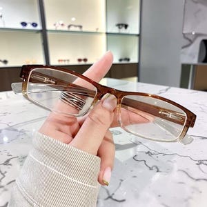 May include: Brown and clear rectangular eyeglasses with a gradient effect. The frames are a dark brown at the top, fading to clear at the bottom. The glasses are held up by a hand, with a marble surface in the background.