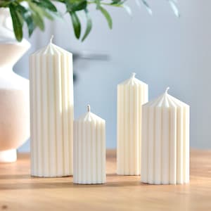 Peut inclure: Quatre bougies piliers ivoire de différentes hauteurs, disposées sur une surface en bois. Chaque bougie présente un design côtelé et un sommet pointu avec une mèche. Les bougies sont de couleur neutre, adaptées à la décoration intérieure.