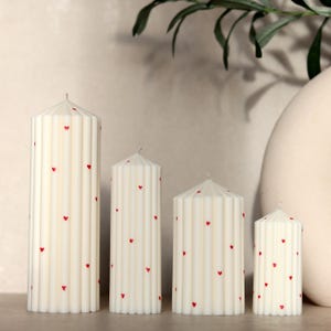 Peut inclure: Quatre bougies piliers ivoire de différentes hauteurs, ornées de petits cœurs rouges. Les bougies ont une texture côtelée et sont disposées en rangée. Ces bougies décoratives sont un élément romantique et décoratif.