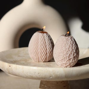 Peut inclure: Deux bougies en forme d'œuf de couleur beige doux, chacune présentant un motif texturé distinct. Une bougie affiche un motif à chevrons vertical, tandis que l'autre présente une grille triangulaire géométrique. Les deux sont allumées.