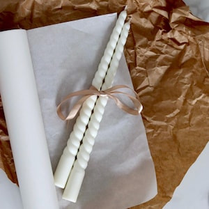 Może przedstawiać: Dwie białe, skręcone świece stożkowe związane jasnobrązową wstążką. Świece leżą na białym papierze, z rolką białego papieru i brązowym papierem w tle. Świece mają spiralny wzór.