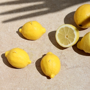 Peut inclure: Trois bougies jaune vif en forme de citron avec de petites mèches blanches, disposées à côté d'un citron entier et d'un demi-citron montrant la pulpe et les segments intérieurs, avec une écorce texturée et réaliste.