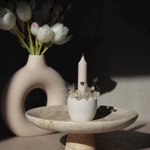Pode incluir: Uma vela bege com um desenho de coração está em um porta-velas branco em forma de casca de ovo, decorado com flores secas e detalhes delicados. A vela está em um pedestal de pedra com um topo redondo, ao lado de um vaso bege com tulipas brancas.