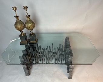 Puede incluir: Una mesa de centro moderna con una tapa de cristal transparente y una base metálica texturizada única. Dos lámparas doradas con detalles ornamentados están sobre la mesa. Las lámparas miden unos 50 cm de altura.