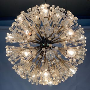 Op de afbeelding: Een bolvormige kroonluchter met meerdere gloeilampen en heldere glazen accenten. De armatuur heeft een centrale kern met uitstralende armen, elk met een lamp. Het ontwerp omvat heldere glazen kralen en bloemvormige glaselementen.