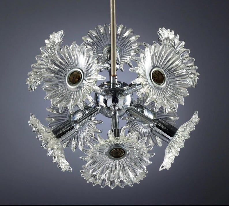 Puede incluir: Un candelabro vintage con pantallas de vidrio transparente en forma de flor. La l&aacute;mpara tiene un cubo central y brazos cromados, con m&uacute;ltiples portal&aacute;mparas. El dise&ntilde;o recuerda la est&eacute;tica moderna de mediados de siglo, ofreciendo una soluci&oacute;n de iluminaci&oacute;n &uacute;nica.