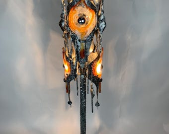 Puede incluir: Una lámpara de pie ornamentada de mediados de siglo. La lámpara presenta un marco de metal oscuro con un centro de vidrio naranja y transparente en forma de ojo. La lámpara tiene una base texturizada y una serie de detalles metálicos colgantes. La lámpara está encendida, emitiendo un brillo cálido.