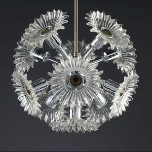 Puede incluir: Una l&aacute;mpara de ara&ntilde;a de cristal transparente y cromo con un dise&ntilde;o esf&eacute;rico. La l&aacute;mpara presenta m&uacute;ltiples pantallas de cristal transparente en forma de flor y brazos cromados, suspendidos de una varilla central. Las luminarias est&aacute;n dispuestas en un patr&oacute;n radial.