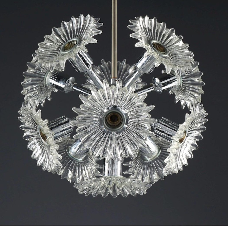 Puede incluir: Una l&aacute;mpara de ara&ntilde;a de cristal transparente y cromo con un dise&ntilde;o esf&eacute;rico. La l&aacute;mpara presenta m&uacute;ltiples pantallas de cristal transparente en forma de flor y brazos cromados, suspendidos de una varilla central. Las luminarias est&aacute;n dispuestas en un patr&oacute;n radial.