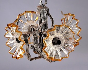 Puede incluir: Una lámpara de araña vintage con una estructura de metal plateado y tres pantallas de cristal con forma de flor. Las pantallas de cristal tienen un centro transparente y bordes de color ámbar. La lámpara cuelga de una cadena.