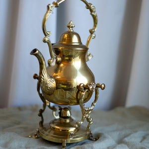 Könnte beinhalten: Eine Vintage-Teekanne aus Messing mit dekorativem Griff und verziertem Sockel. Die Teekanne hat einen gebogenen Ausguss und einen Deckel mit einem kleinen Knauf. Das Messing hat eine polierte, reflektierende Oberfläche. Die Teekanne steht auf einem passenden Ständer.