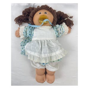May include: A vintage Cabbage Patch Kid doll with brown yarn hair styled in pigtails. The doll has a pacifier and is wearing a blue floral print dress with a white eyelet trim and matching bloomers. The doll's skin is a peach color.