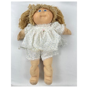 May include: A vintage doll with a soft, fabric body and a vinyl face. The doll has blonde yarn hair, blue eyes, and is wearing a white outfit with blue leaf patterns and lace trim. A small pink flower is on the chest.