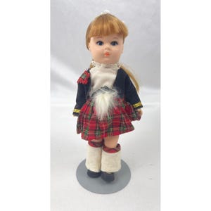 May include: A vintage doll dressed in traditional Scottish attire. The doll has red hair, blue eyes, and is wearing a black jacket with gold trim, a red plaid kilt, white socks, and black shoes. The doll is standing on a gray base.
