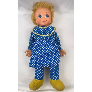 May include: A vintage doll with a smiling face, blue eyes, and blonde hair. The doll wears a blue dress and pants with white polka dots, a yellow-trimmed collar, and yellow shoes. The doll is a classic toy.