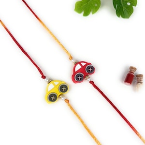 May include: Two decorative wristbands featuring car-shaped charms. One charm is yellow and the other is red, both with black wheel details. The charms are attached to thin, vibrant red and orange threads accented with small white beads.
