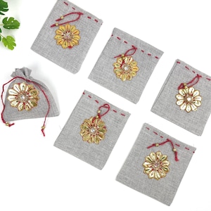 May include: A collection of gray linen drawstring bags, each adorned with a gold and red floral embellishment. The bags are arranged on a white surface, showcasing their decorative design and potential use for small gifts or favors.
